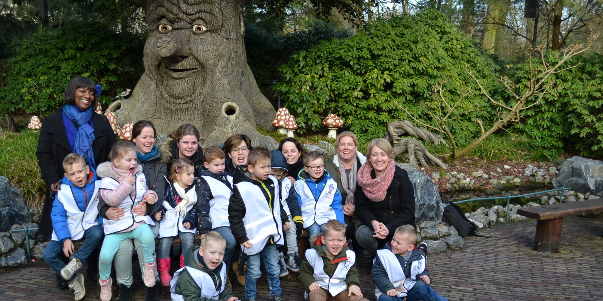 A group of children and adults posing in front of a large tree sculpture with a friendly face in a themed park.