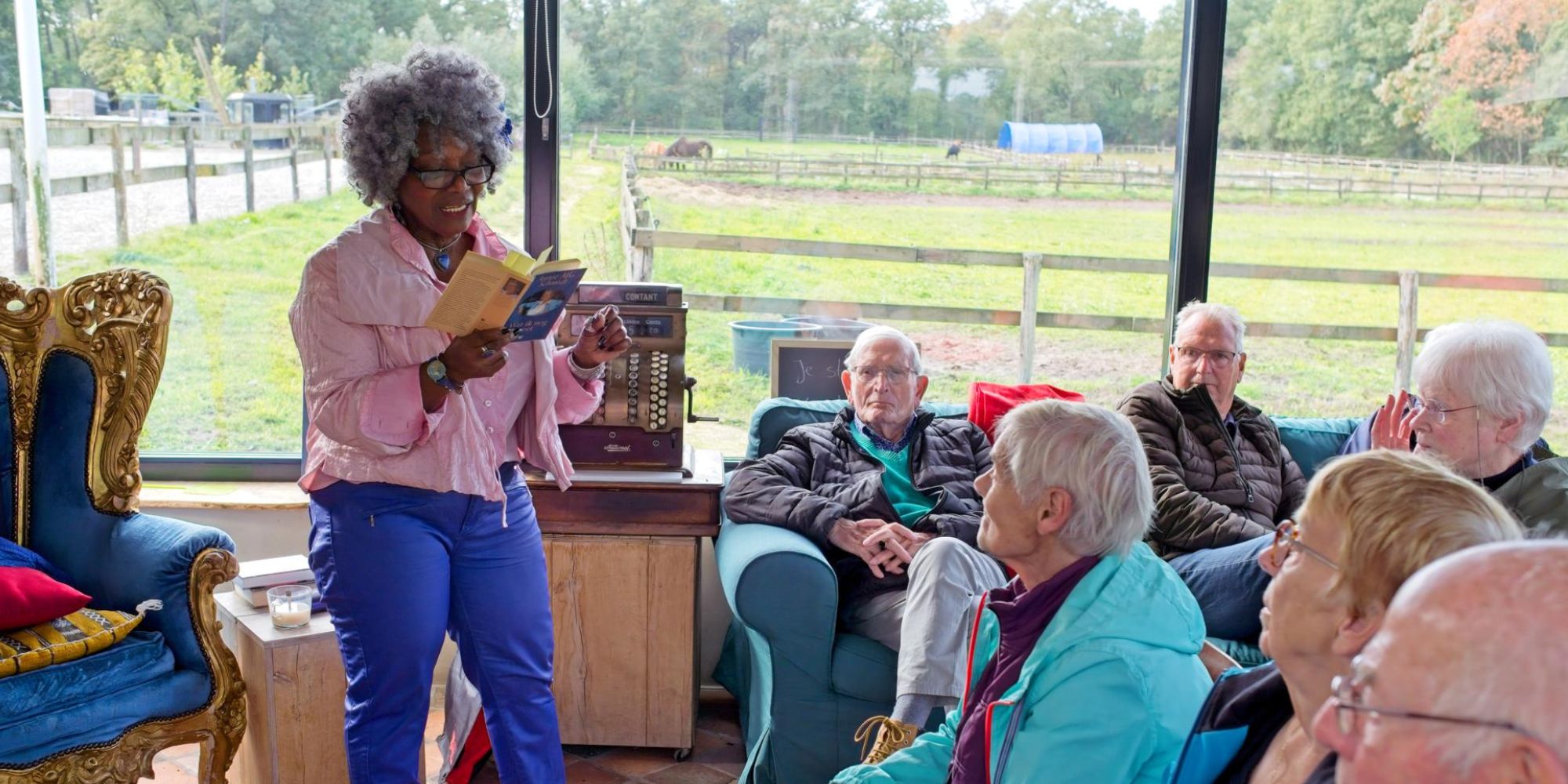 A woman with gray hair and glasses reading a book to a group of elderly people in a cozy rural setting with a farm in the background.