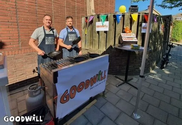 Two men grilling pancakes at an outdoor event under a "GOODWILL" banner, with colorful decorations around.