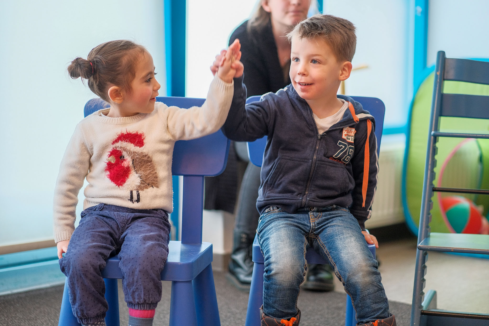 A young boy and girl sitting on blue chairs, smiling and high-fiving each other in a classroom.