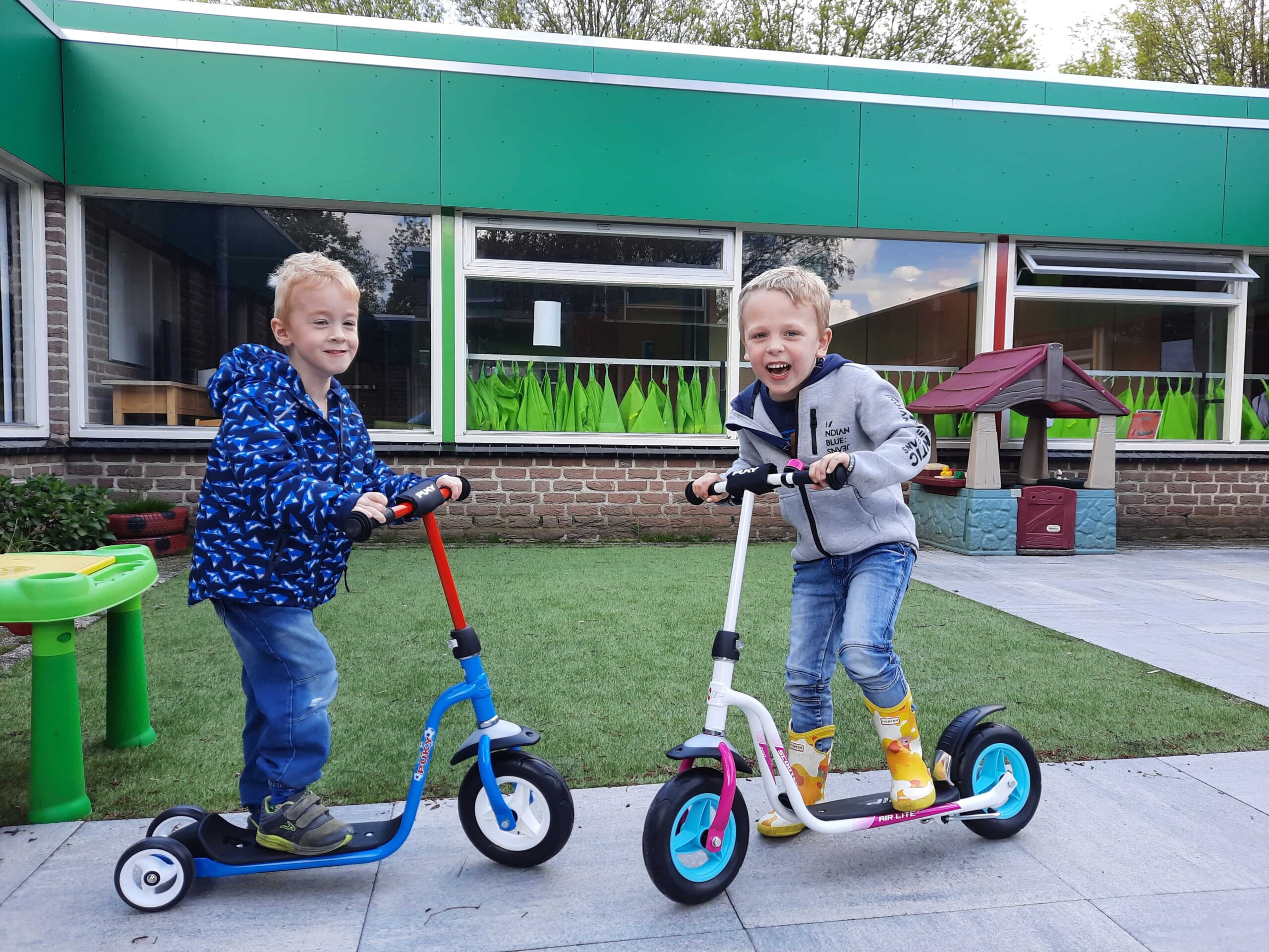 Two young boys riding scooters on a paved area outside a school or daycare.