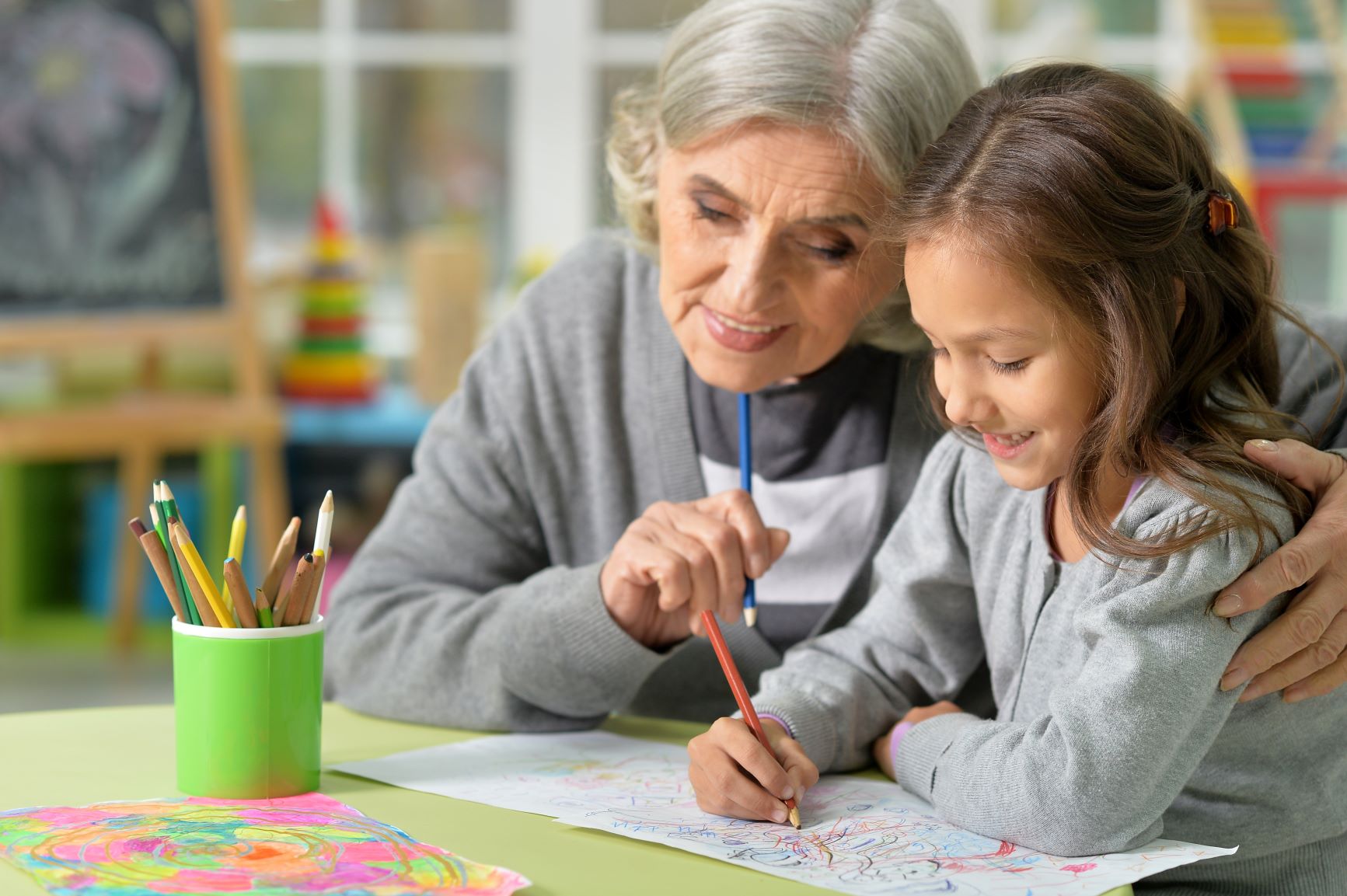 An elderly woman and a young girl happily drawing together at a table in a classroom.