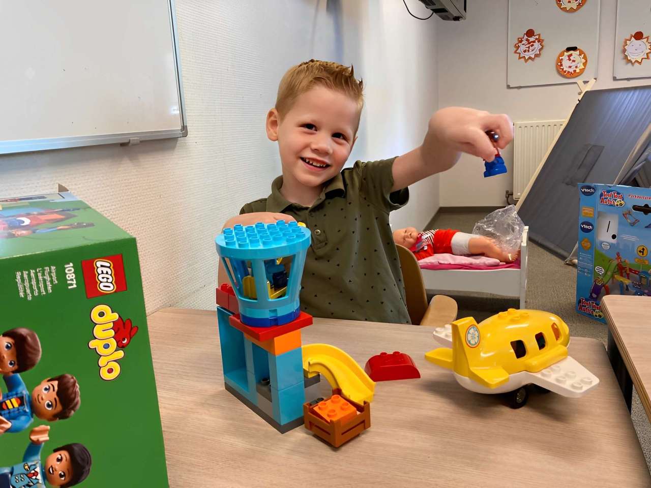 A young boy playing with LEGO Duplo toys, smiling at the camera in a classroom setting.