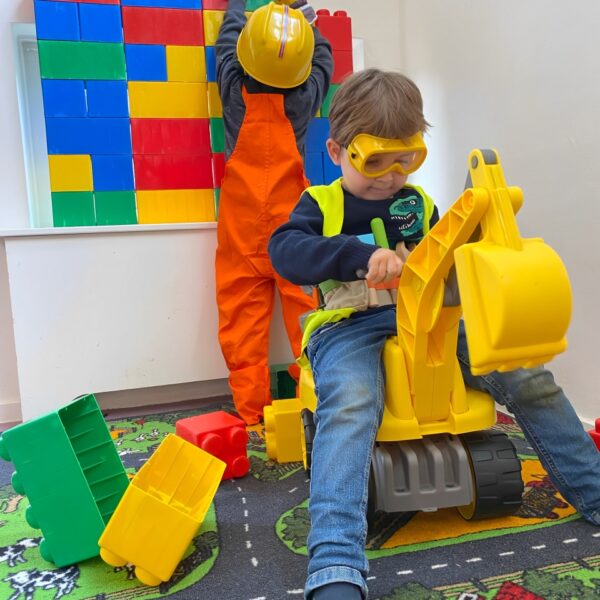 Two children dressed as construction workers playing with large toy bricks and a toy excavator.