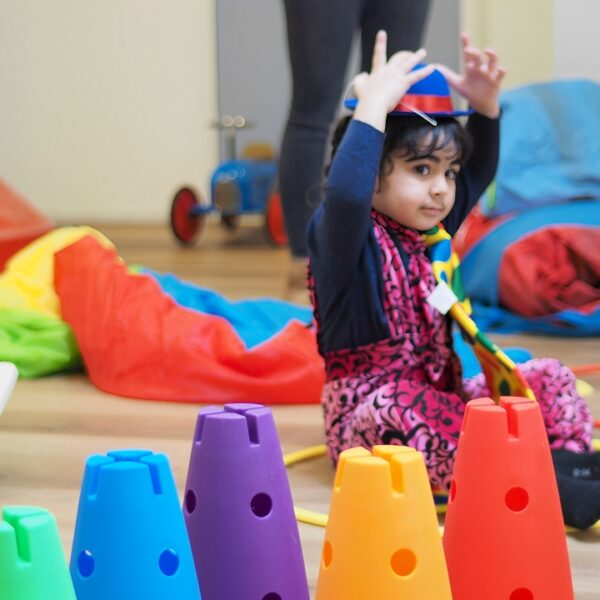A young girl sitting on the floor playing with colorful cones, hoops, and other toys indoors.