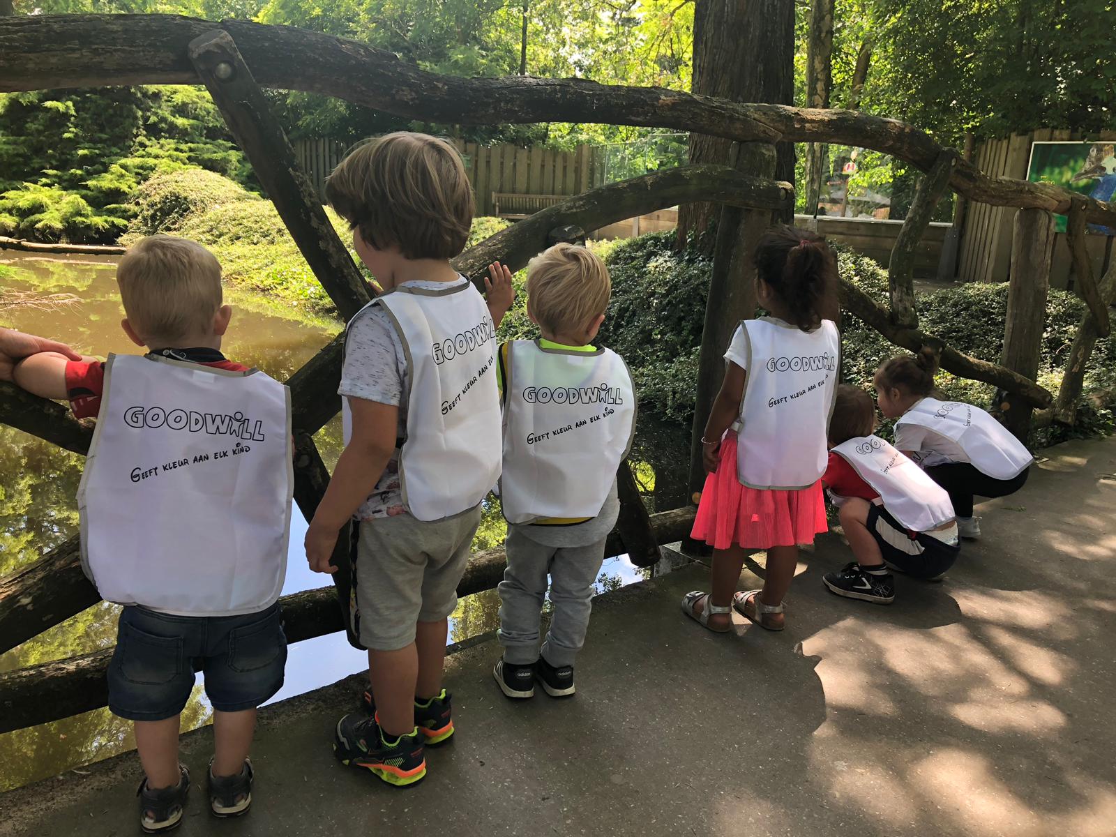 A group of young children wearing white vests with "GOODWILL" written on the back, standing near a wooden fence looking at a pond.
