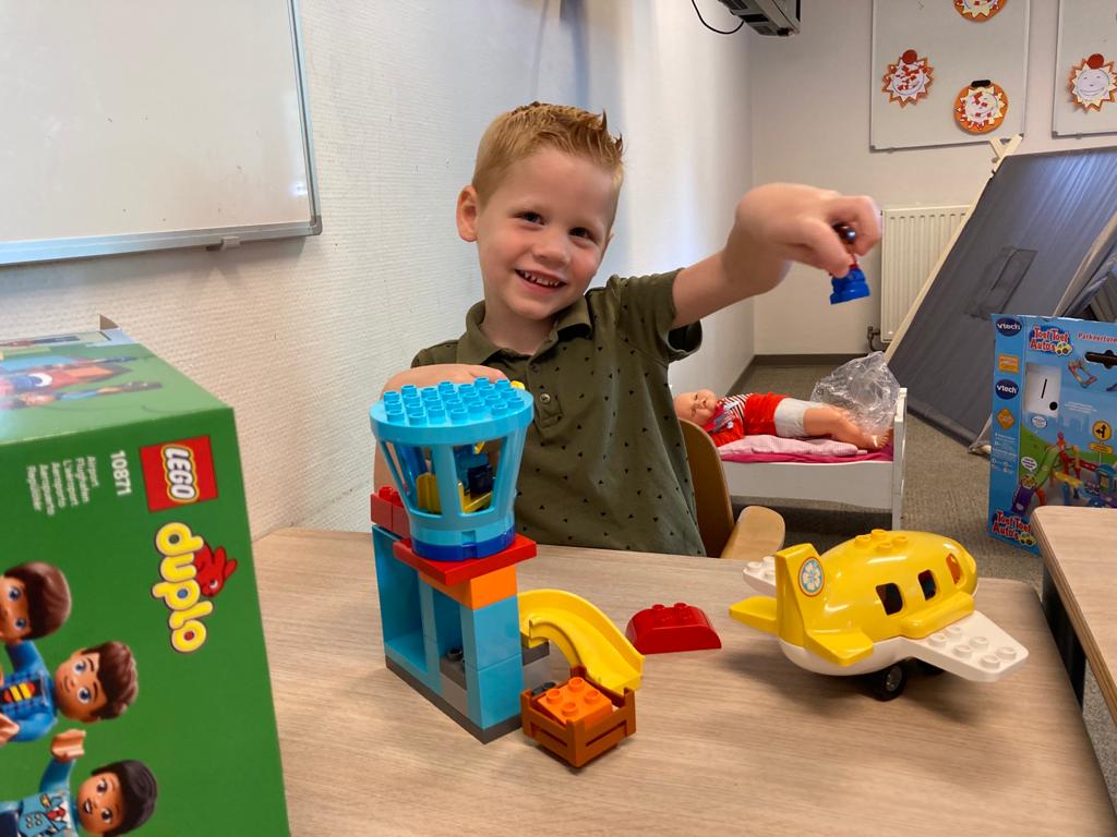 A young boy sitting at a table, playing with LEGO Duplo and toy airplane, smiling at the camera.