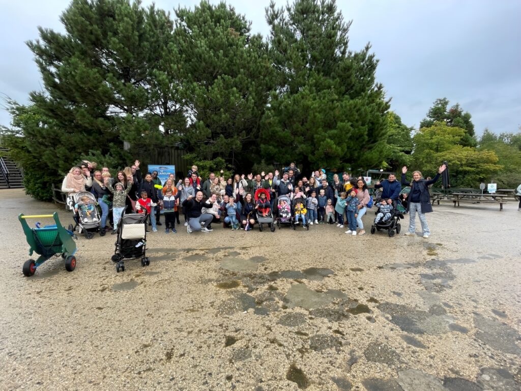 A large group of families with children in strollers, gathered outdoors, waving at the camera.