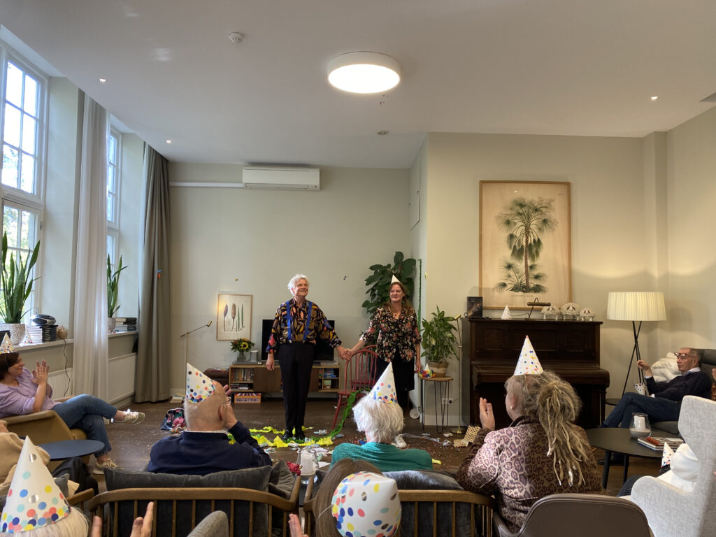 A cozy indoor gathering with elderly people wearing party hats, watching two women standing at the front of the room.