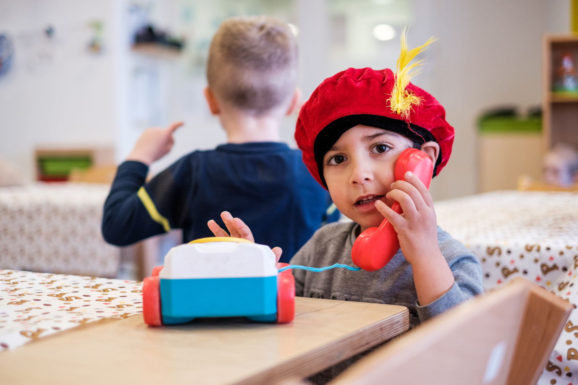 A young child wearing a red hat with a feather, playing with a toy telephone while sitting at a table.