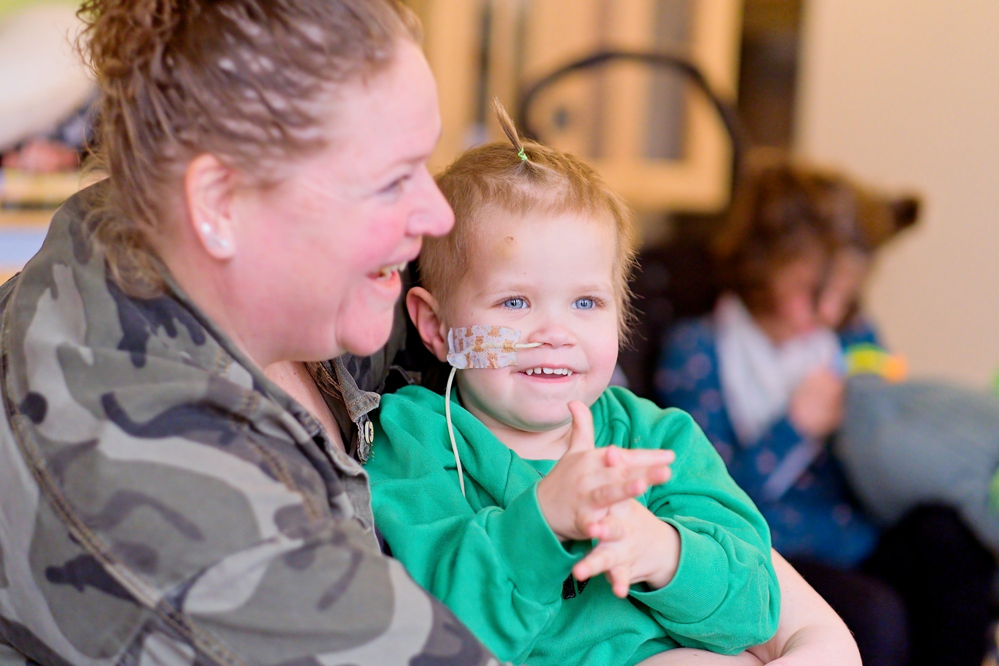 A woman in camouflage clothing smiling while holding a young child with a nasal tube, both looking joyful.